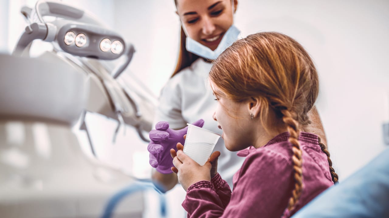 A girl sits up in a dentists chair drinking water which has been given to her by the dental nurse.