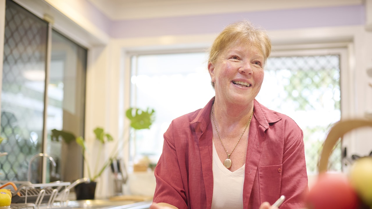 A middle-aged woman stands at a kitchen bench holding a bowl and fork and smiling.