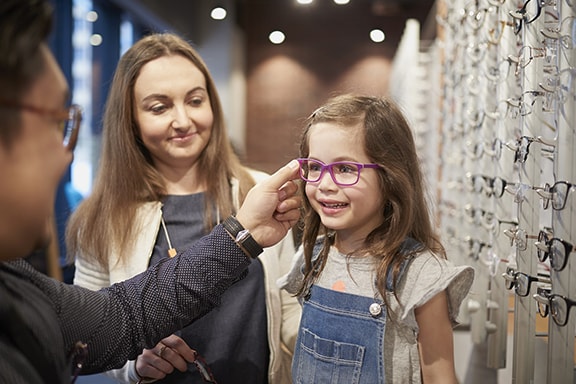 A child wearing glasses, standing with a woman and an optometrist who is checking her glasses.