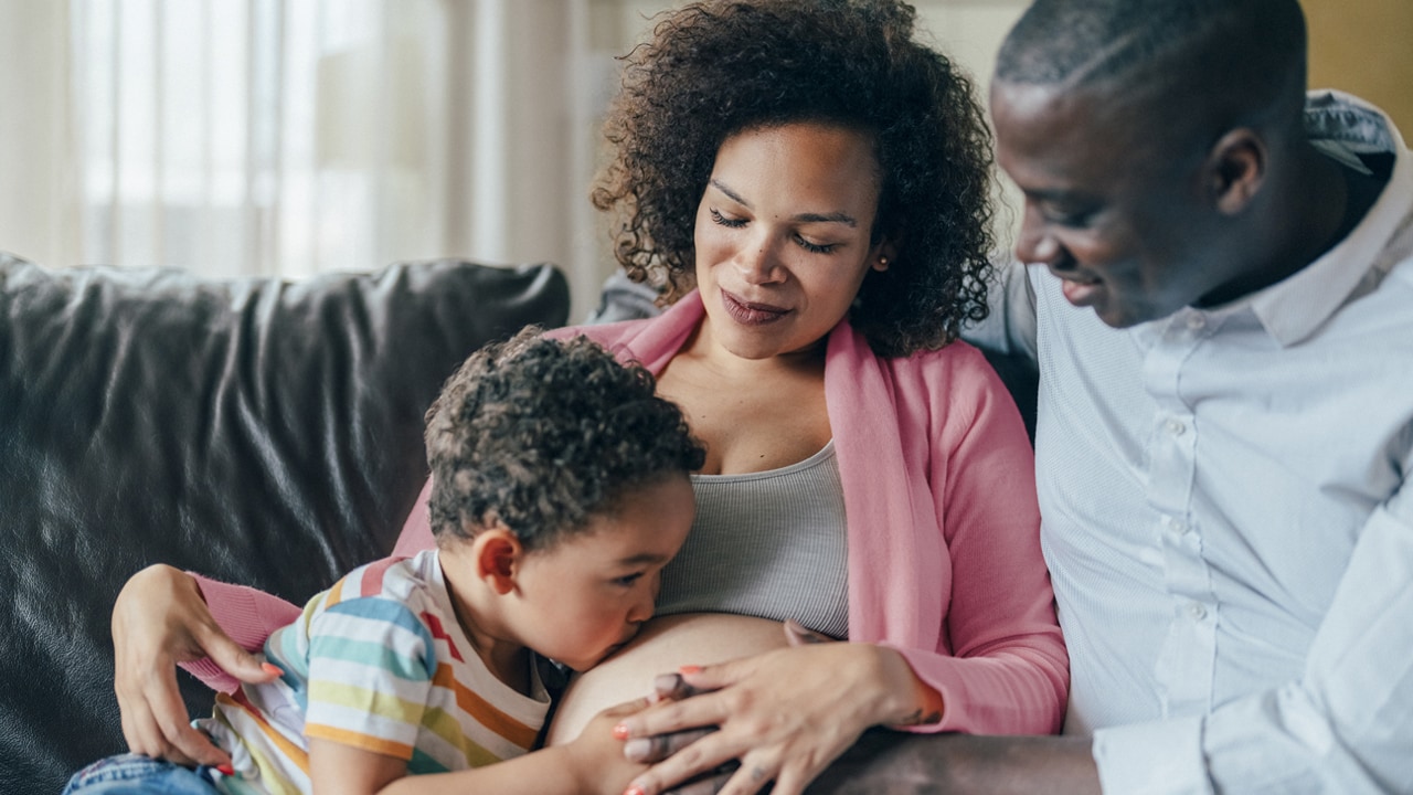 A pregnant mother sits on the couch with her partner and child who kisses her pregnant belly.