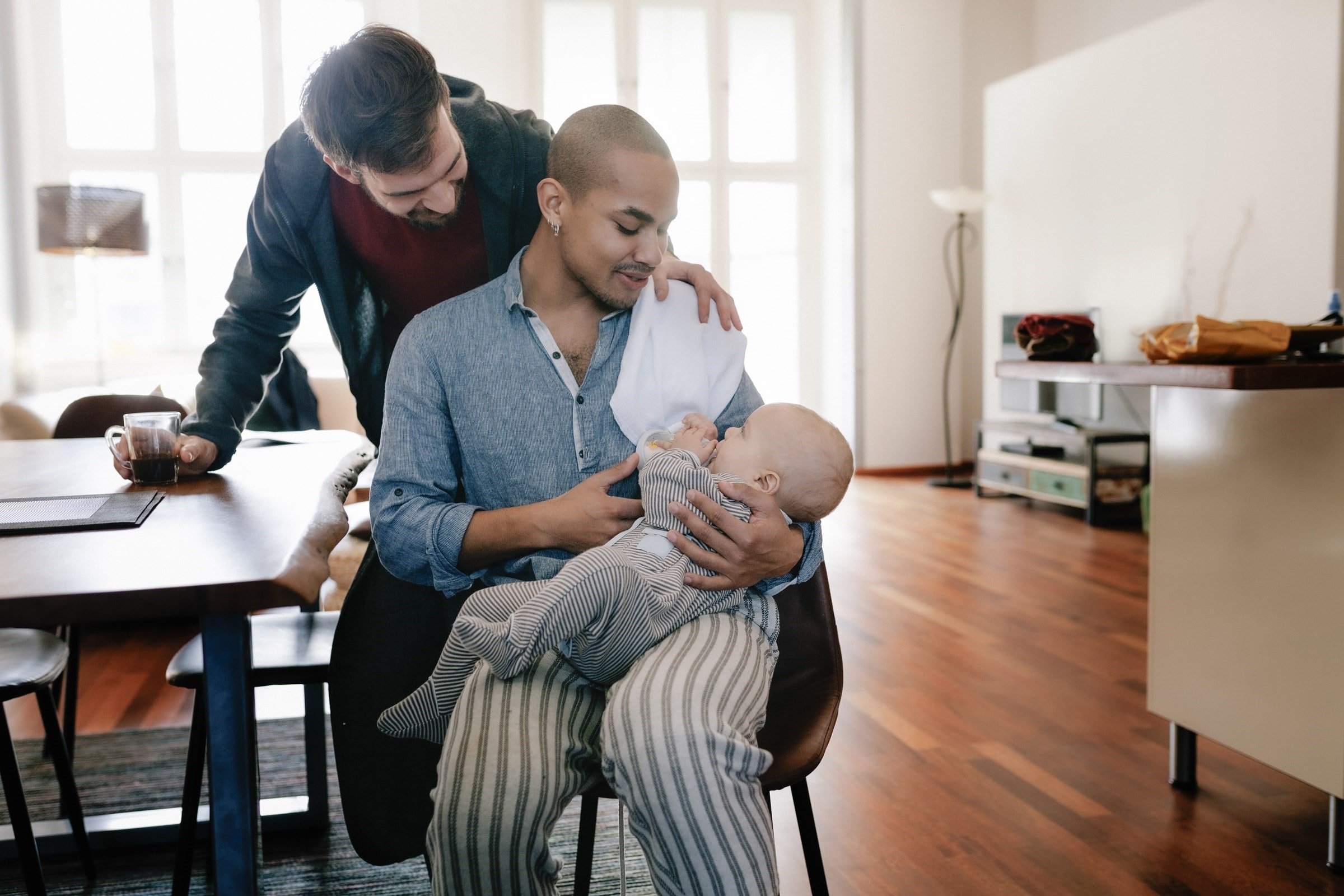 A man sits at a table feeding a baby with a bottle while his male partner holds his shoulder from behind.