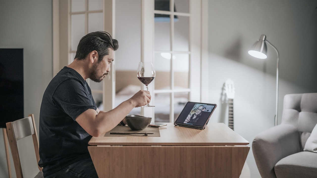 A man sits alone at a table in his home and eats dinner while drinking red wine and watching a video.