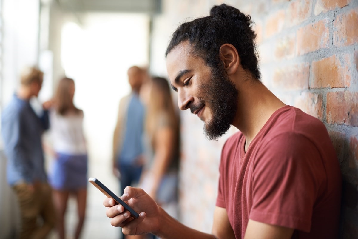 A man stands with his back against a brick wall while looking at his phone and smiling.