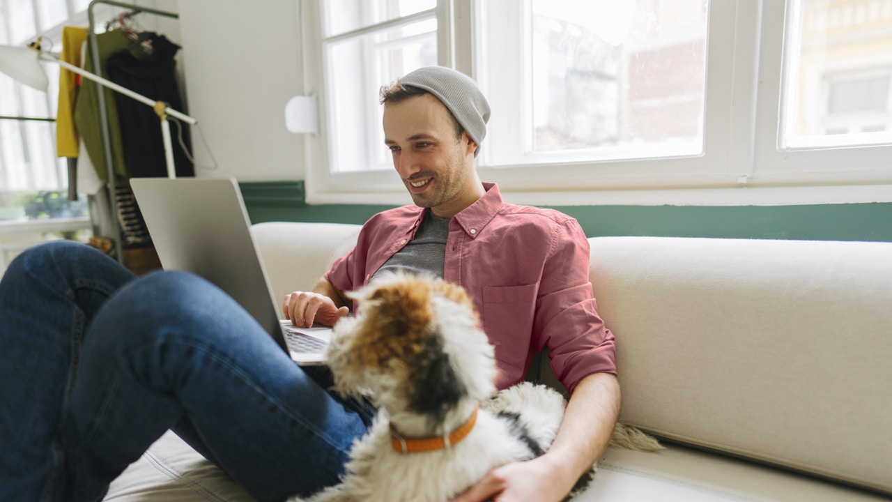  A young man sits on a couch with his dog next to him and a laptop open on his lap.