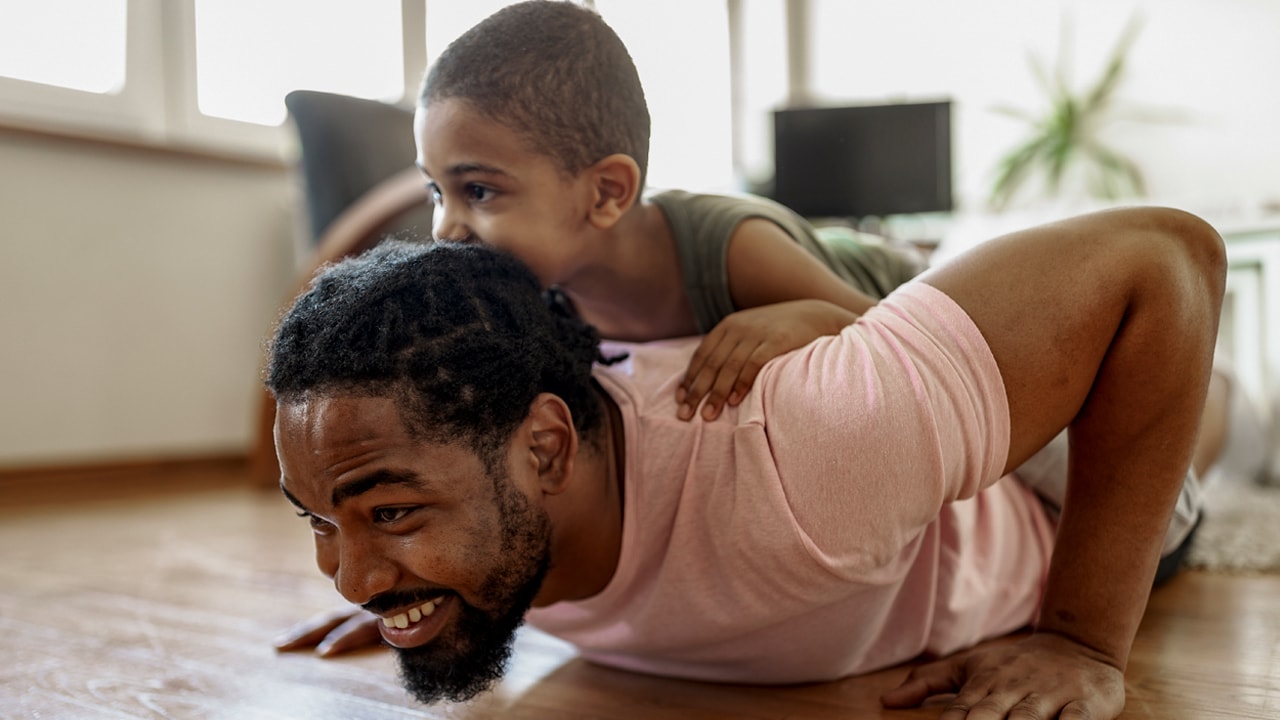  A man does push-ups on the floor in his home and his young son lies on his back.