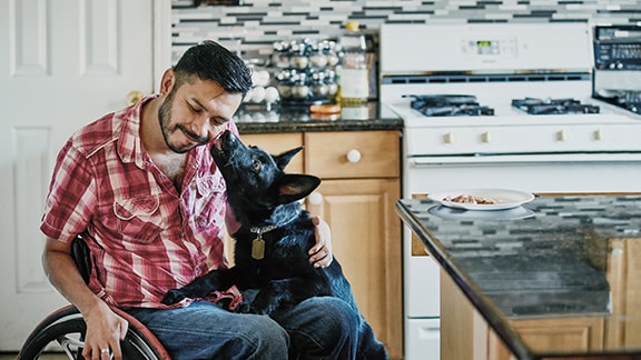 A man sitting in a wheelchair in a kitchen with a dog licking his face