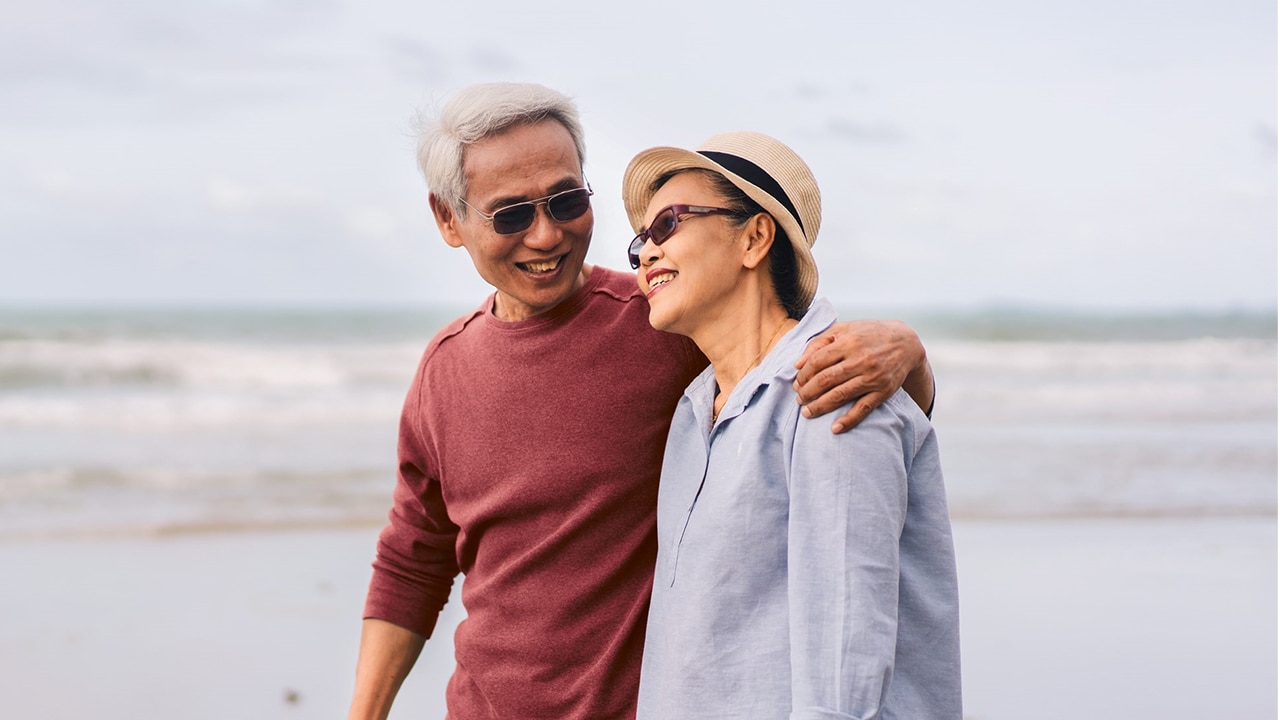 A middle aged man and woman walk on a beach, smiling at each other. The man's arm is around the woman's shoulder.