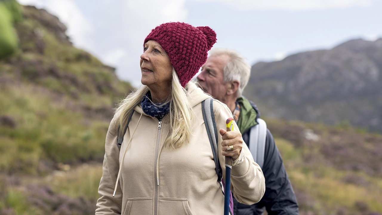 A middle aged couple hike with walking poles, hills behind them.