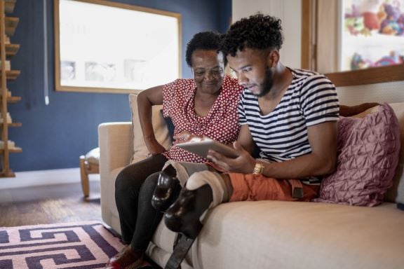 A young man with prosthetic legs, sitting on a couch with a tablet on his lap, showing his grandmother something on the screen.