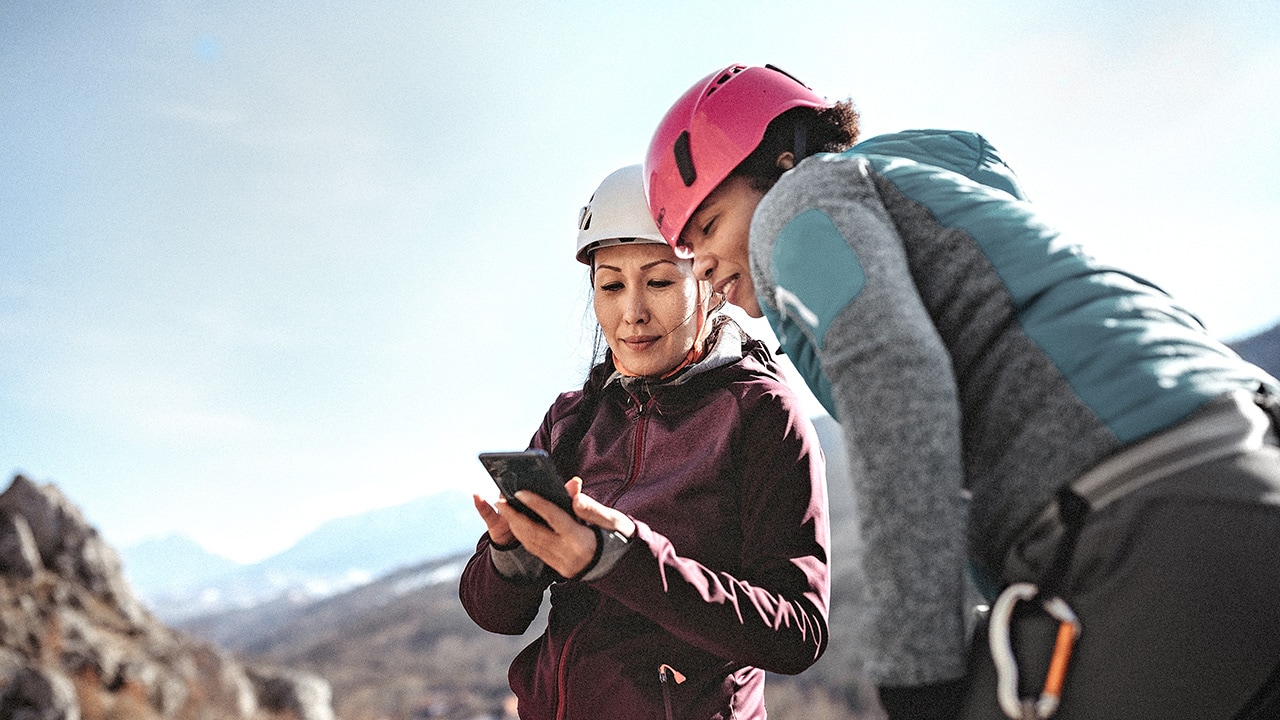 Two female mountaineers with helmets and climbing gear stand high on a mountain and look at a phone.