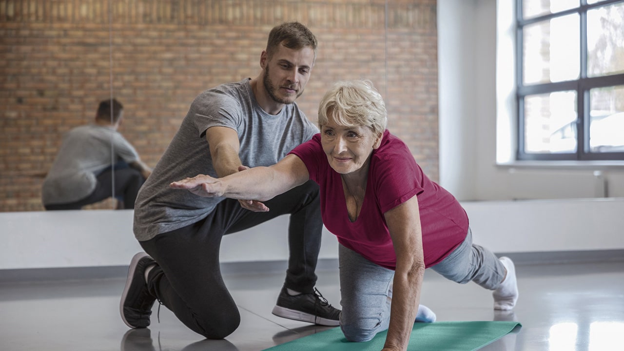 A senior woman does a yoga pose on one hand and one knee with the help of a male trainer.