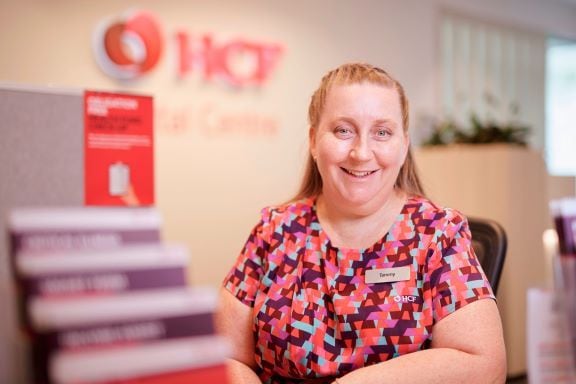 A woman wearing a HCF health insurance company t-shirt with nametag, sitting at a desk and smiling to the camera.