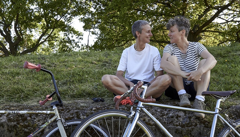 Two women sit close on a hill, their bikes on the ground in front of them. They smile at each other.