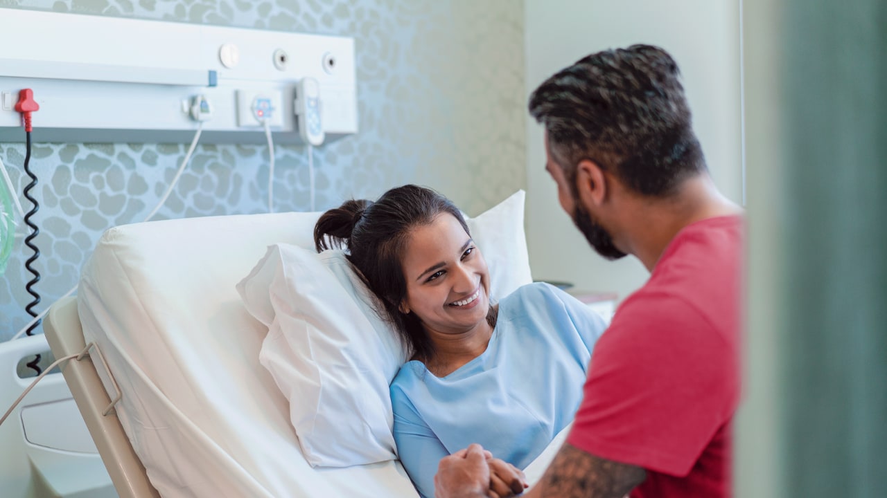  A woman lies in a hospital bed and smiles at her male partner who sits on the edge of the bed looking at her.