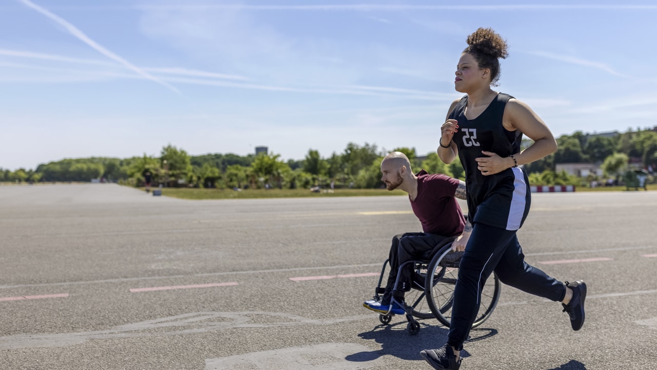 A man runs on a road and another man in a wheelchair rolls alongside him.