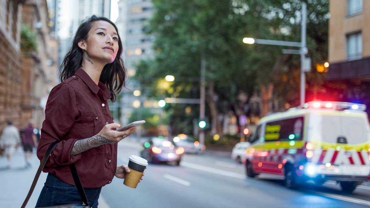 A woman stands on a footpath in the city as an ambulance drives past in the background with flashing lights, she holds a phone and looks down the street.