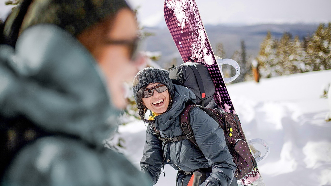 Two women hike a snowy hill with snowboards strapped to their backs and laugh together.